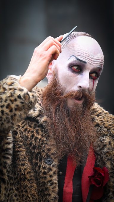 Man med skägg och smink i leopardmönstrad jacka, klipper sitt hår.